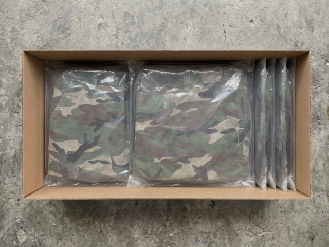 Top-down view of camouflage trousers in clear poly bags, jumbled in a cardboard shipping box on a warehouse floor