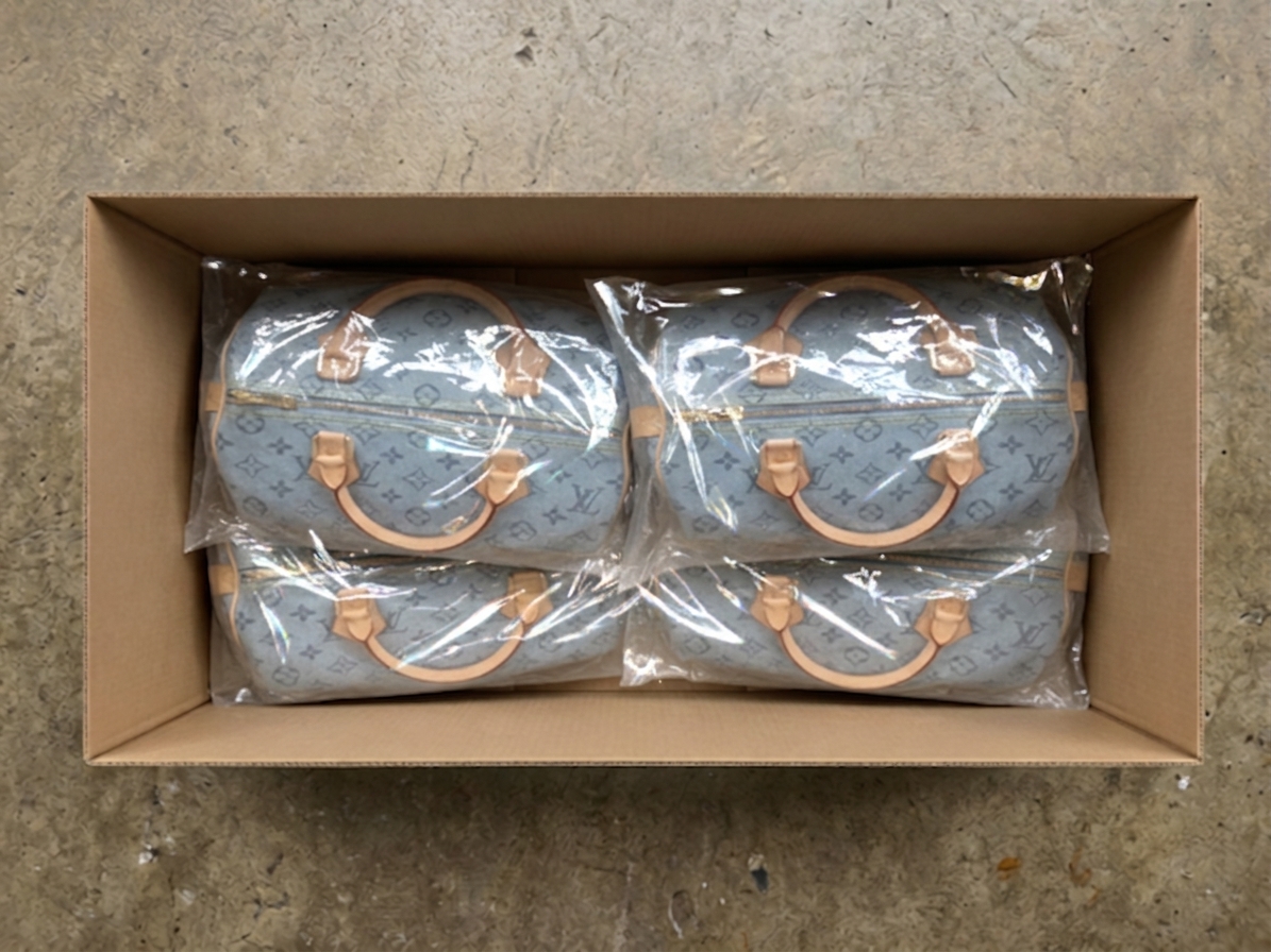 Top-down view of monogram denim handbags in clear poly bags, arranged in a 2×2 grid inside a cardboard box